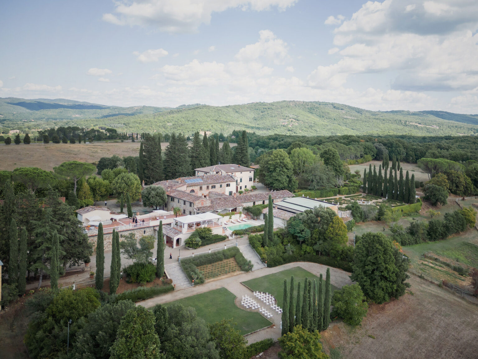 Aerial view of a historic villa surrounded by lush greenery and rolling hills in Tuscany, Italy, featuring manicured gardens, cypress trees, and an outdoor event area, ideal for weddings and retreats. | Only on SopranoVillas