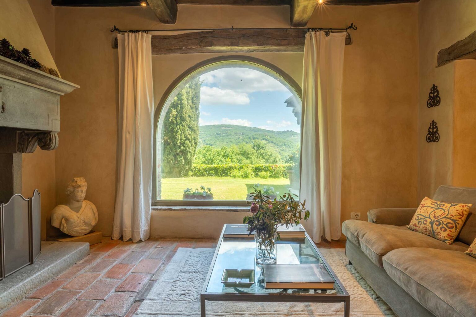 Cozy living room featuring a large arched window with scenic views of rolling hills, natural light streaming in, a comfortable sofa with decorative pillows, a glass coffee table, and an elegant bust sculpture on the side. | Only on SopranoVillas