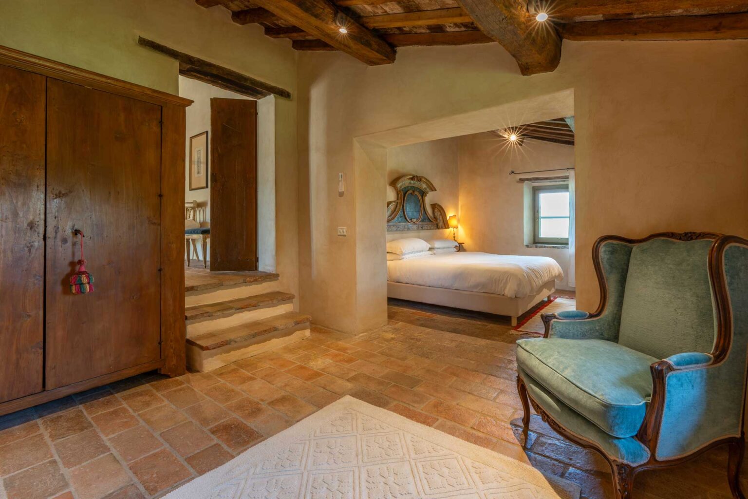 Cozy bedroom interior featuring a blue accent chair, wooden wardrobe, and a staircase leading to a well-lit bedroom with a large bed. Rustic decor with stone flooring and warm lighting creates an inviting atmosphere. Ideal for showcasing a charming vacation rental or home design. | Only on SopranoVillas