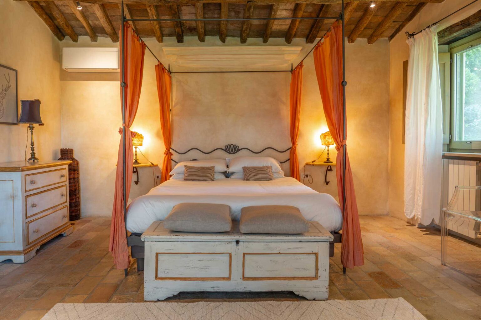 Cozy bedroom featuring a four-poster bed with orange drapes, elegant bedside lamps, and a rustic wooden chest at the foot of the bed, set against a warm, earthy color palette and exposed wooden beams. | Only on SopranoVillas