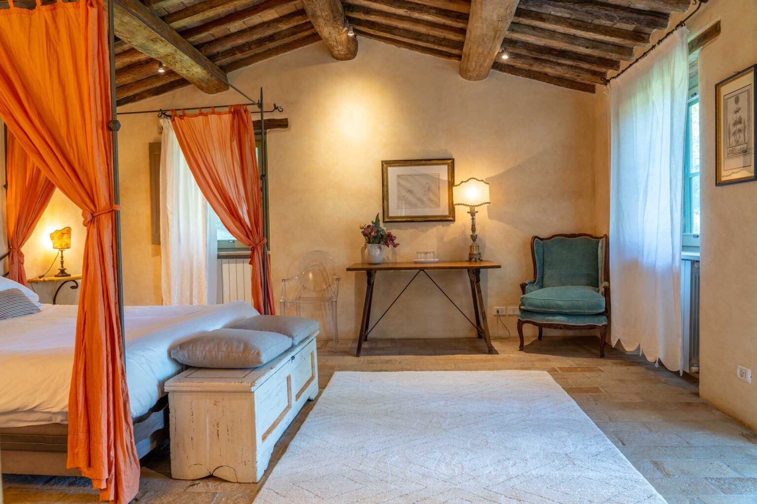 Spacious rustic bedroom featuring a wooden ceiling, orange drapery, a cozy bed with gray pillows, a decorative lamp on a wooden table, and a blue armchair by the window. | Only on SopranoVillas