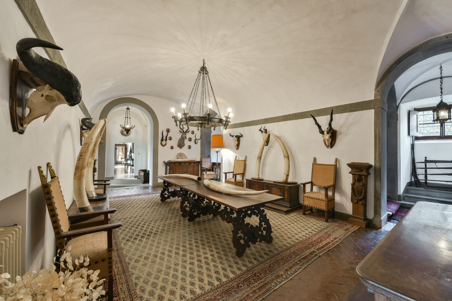 Elegant interior of a vintage dining room featuring a long wooden table, ornate chairs, and decorative taxidermy on the walls, illuminated by a chandelier. The room showcases a blend of rustic and classic design elements. | Only on SopranoVillas