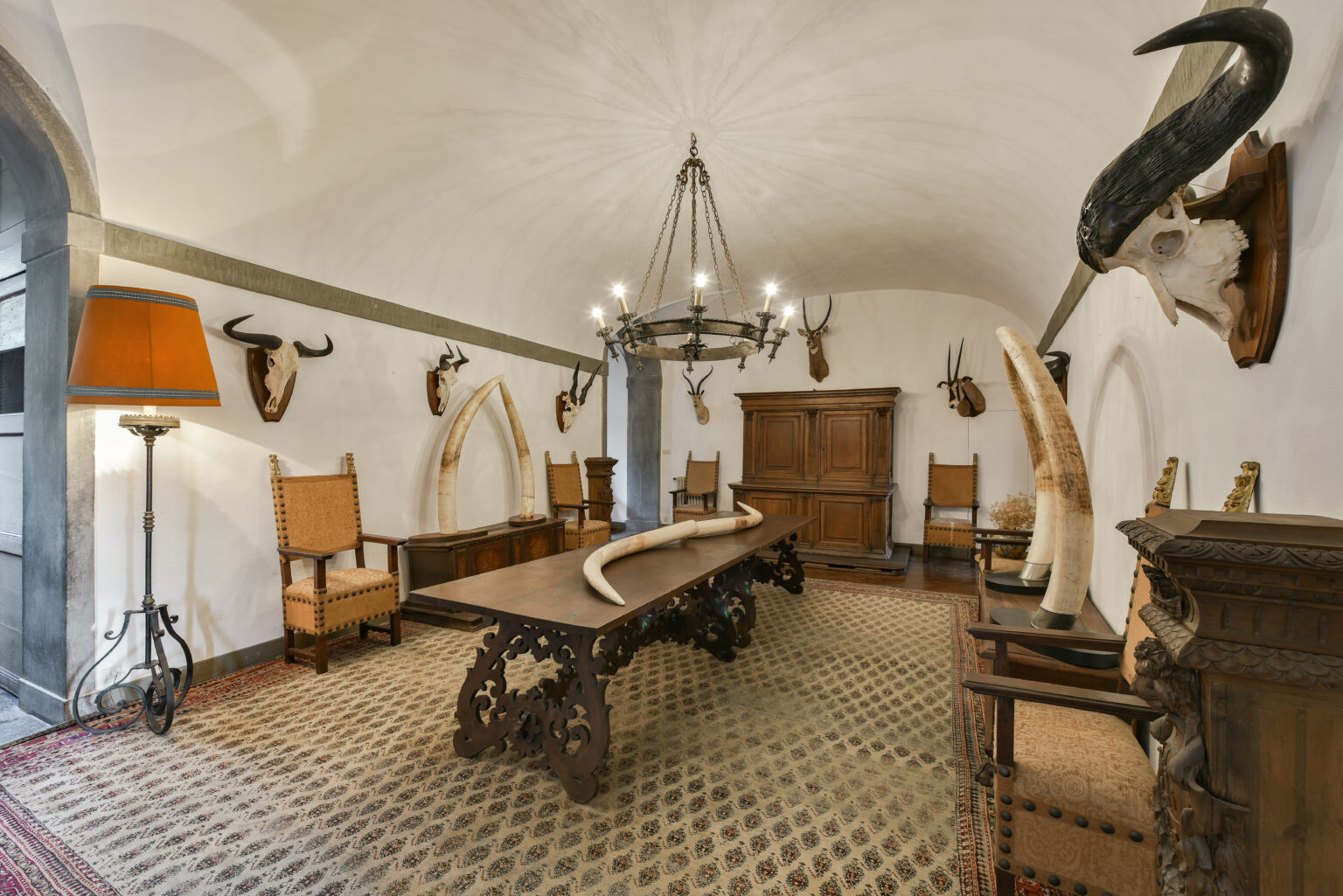 Interior view of a rustic dining room featuring a long wooden table, vintage chairs, and decorative animal horns mounted on the walls. A chandelier hangs from the ceiling, and an ornate wooden cabinet adds to the room's character. The floor is adorned with patterned tiles, creating an inviting atmosphere. | Only on SopranoVillas