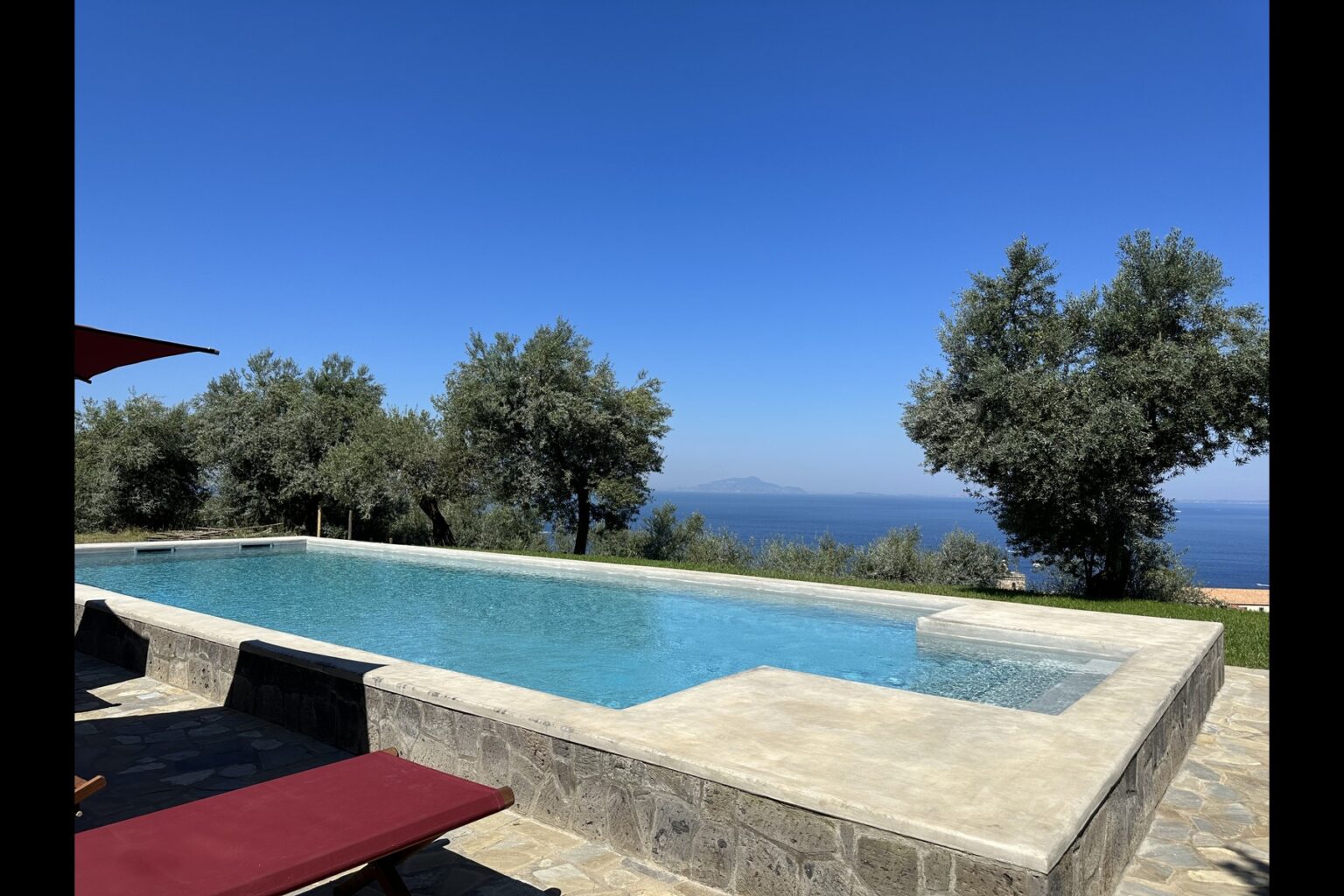Luxurious infinity pool overlooking a clear blue sky and distant ocean, surrounded by olive trees, perfect for relaxation and outdoor living. | Only on SopranoVillas