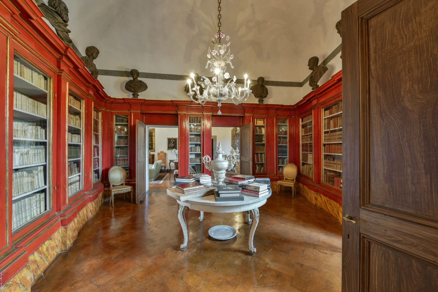 Elegant library interior featuring red-paneled walls, a central round table, and a chandelier, surrounded by shelves filled with books. | Only on SopranoVillas