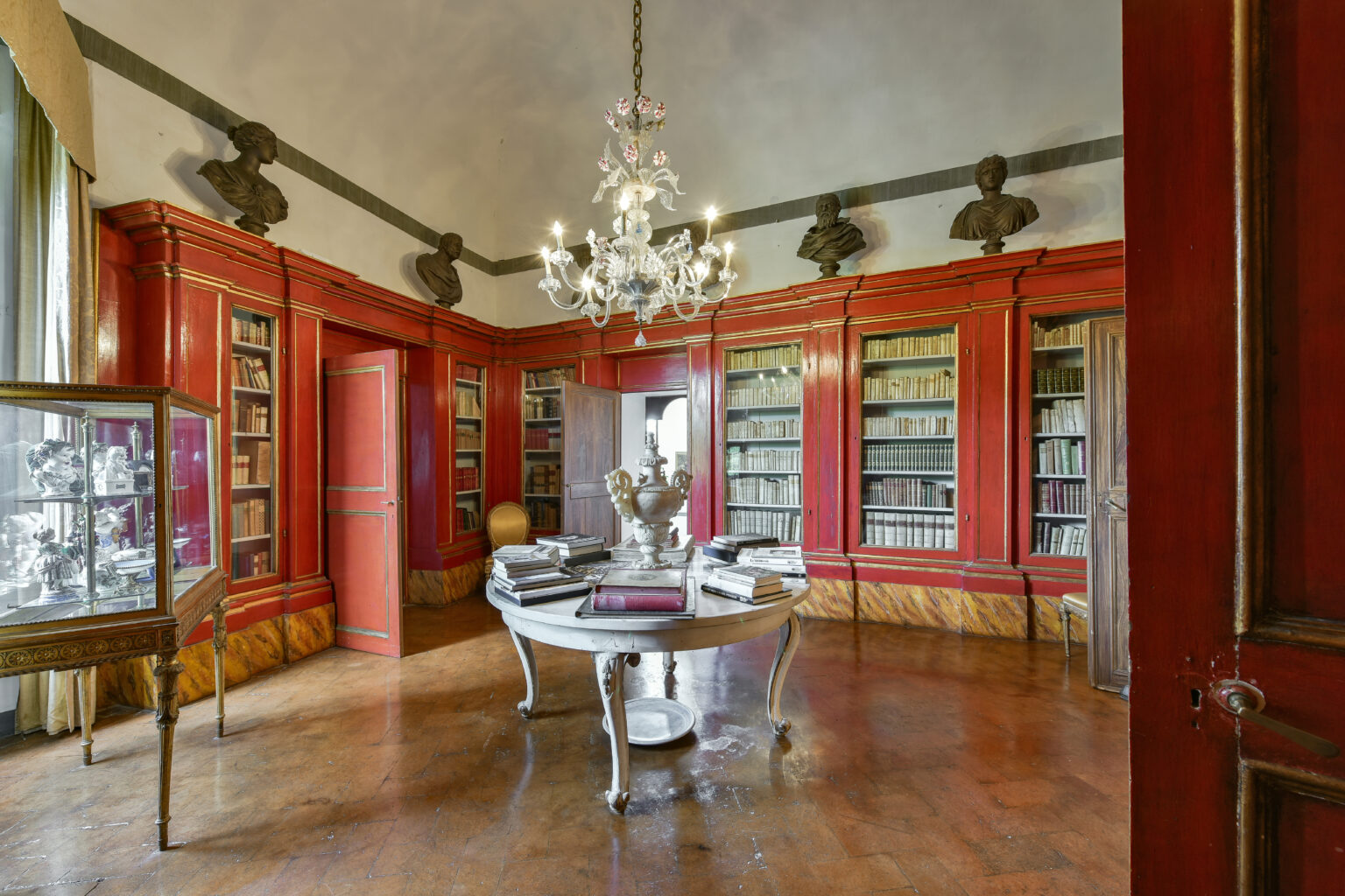 Elegant library interior featuring red wooden bookshelves lined with books, a round table with stacked volumes, and decorative busts on display. A chandelier hangs from the ceiling, adding a touch of sophistication to the warm, inviting atmosphere. | Only on SopranoVillas
