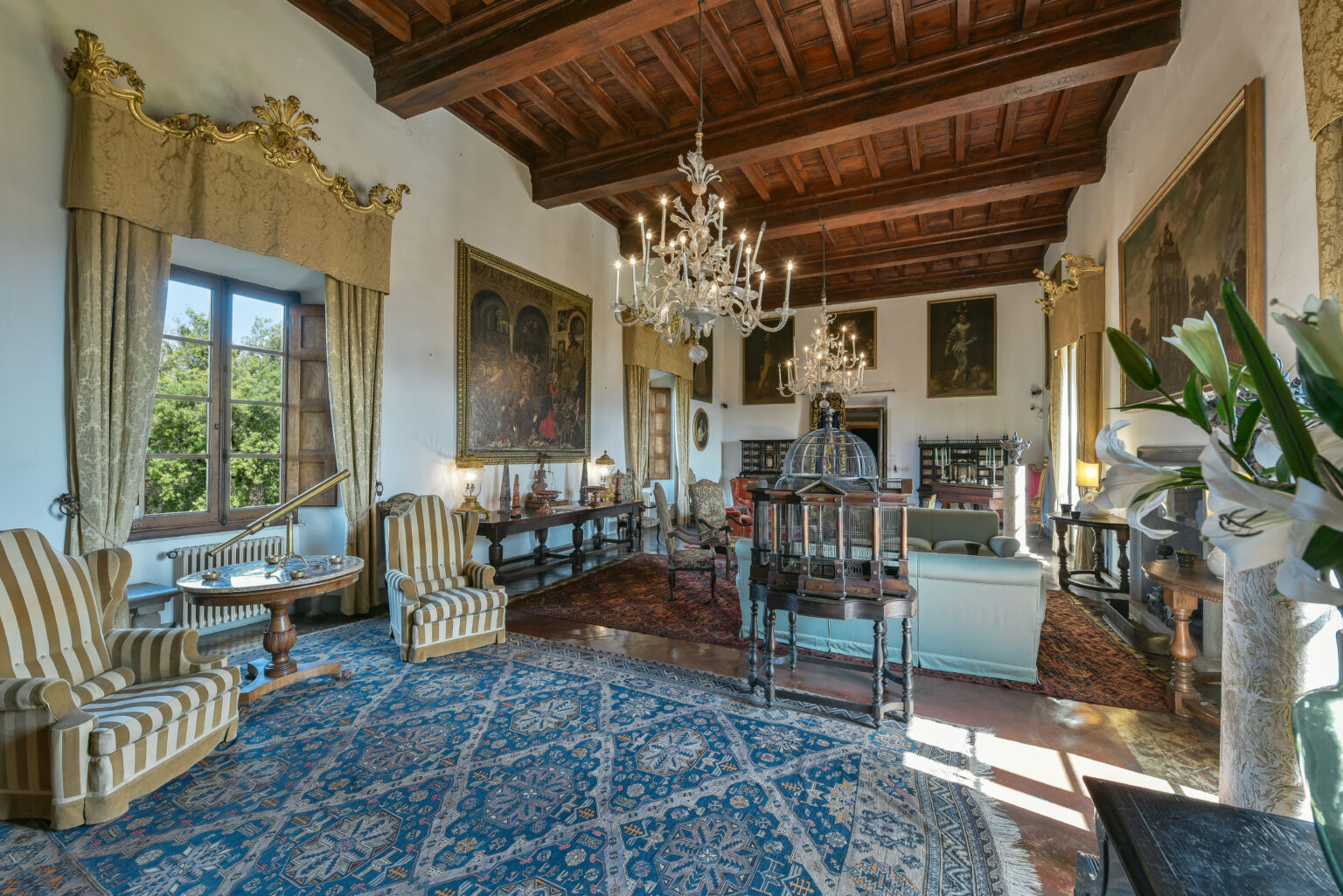 Luxurious living room featuring elegant striped armchairs, intricate chandeliers, and classic artwork, all set against a backdrop of wooden beams and a decorative rug. | Only on SopranoVillas