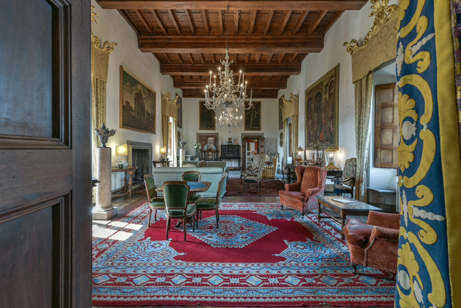 Opulent interior of a historic living room featuring a large chandelier, elegant furniture, and intricate rugs. The room showcases high ceilings with wooden beams and classic artwork on the walls, creating a luxurious atmosphere. | Only on SopranoVillas