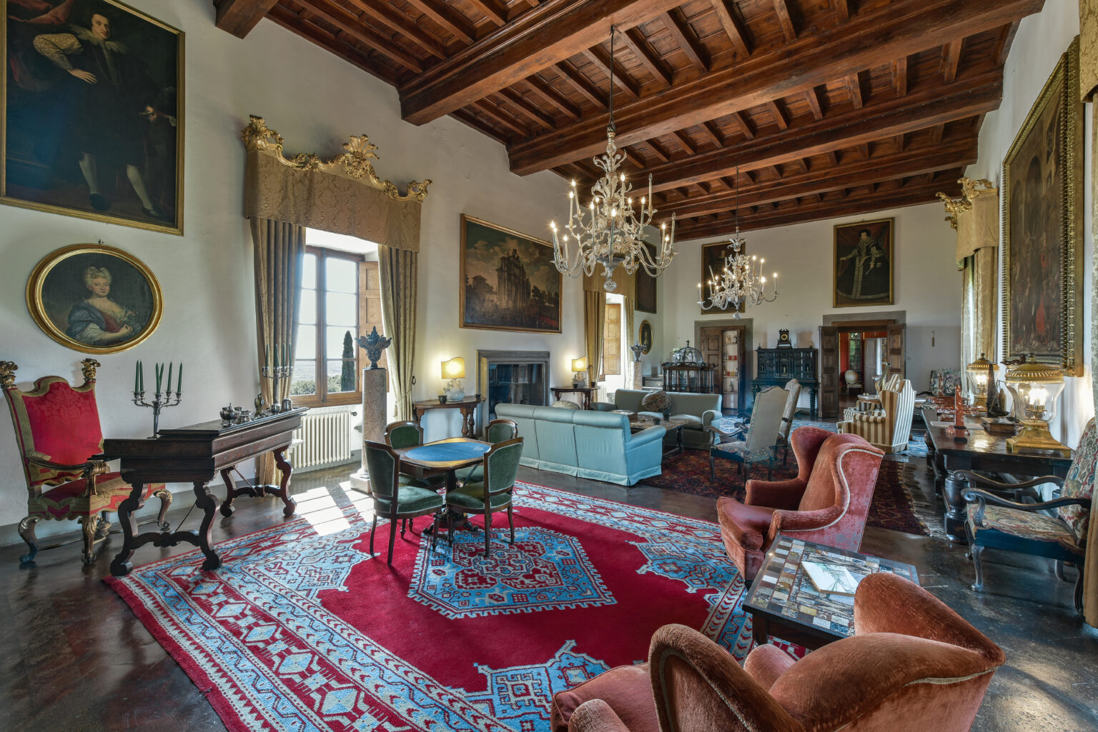 Elegant interior of a historic room featuring a wooden beamed ceiling, ornate chandeliers, and classic furniture. The space includes a vibrant red area rug, paintings on the walls, and a cozy seating arrangement, creating a warm and inviting atmosphere. | Only on SopranoVillas