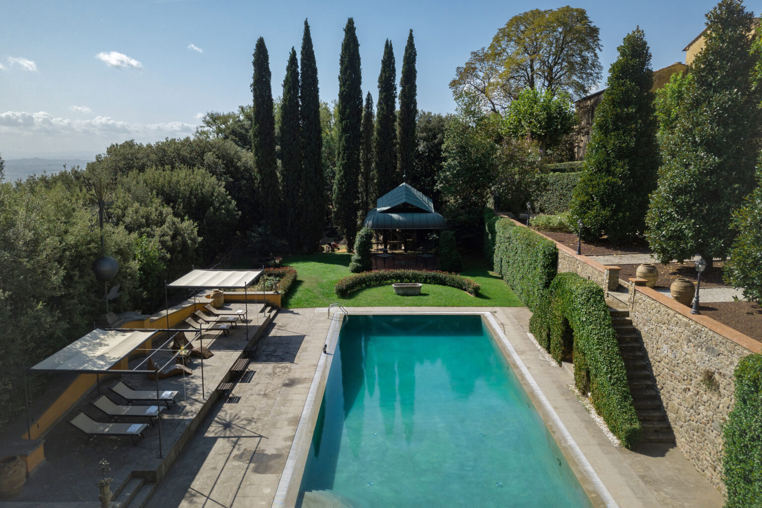 Luxurious outdoor swimming pool surrounded by lush greenery, cypress trees, and a shaded gazebo, offering a serene retreat in a picturesque landscape. | Only on SopranoVillas