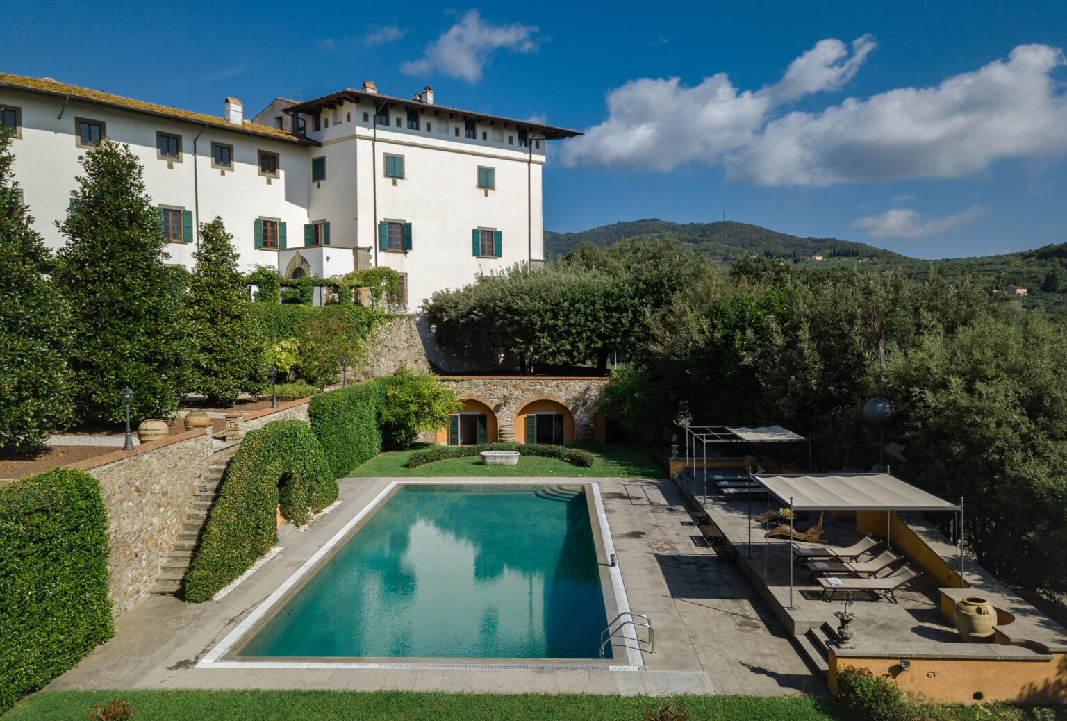 Scenic view of a luxurious villa with a swimming pool, surrounded by lush greenery and olive trees, set against a backdrop of rolling hills and blue skies. | Only on SopranoVillas
