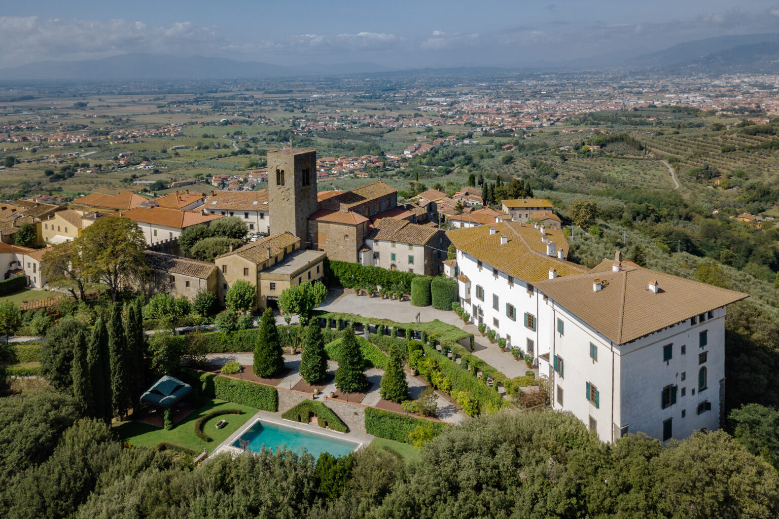Aerial view of a historic Italian villa surrounded by lush greenery, gardens, and a swimming pool, with rolling hills and a distant town in the background. The scene captures the charm of traditional architecture with terracotta roofs and a prominent stone tower. Ideal for showcasing picturesque Tuscan landscapes and luxury accommodations. | Only on SopranoVillas