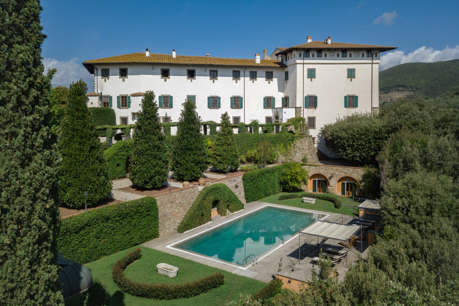 Luxurious villa with a swimming pool surrounded by manicured gardens and trees, set against a scenic hillside backdrop. Ideal for relaxation and upscale retreats. | Only on SopranoVillas
