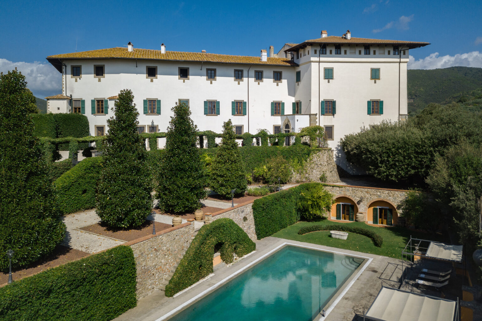 Aerial view of a luxurious villa surrounded by manicured gardens and tall trees, featuring a swimming pool in the foreground and a scenic mountain backdrop under a clear blue sky. | Only on SopranoVillas