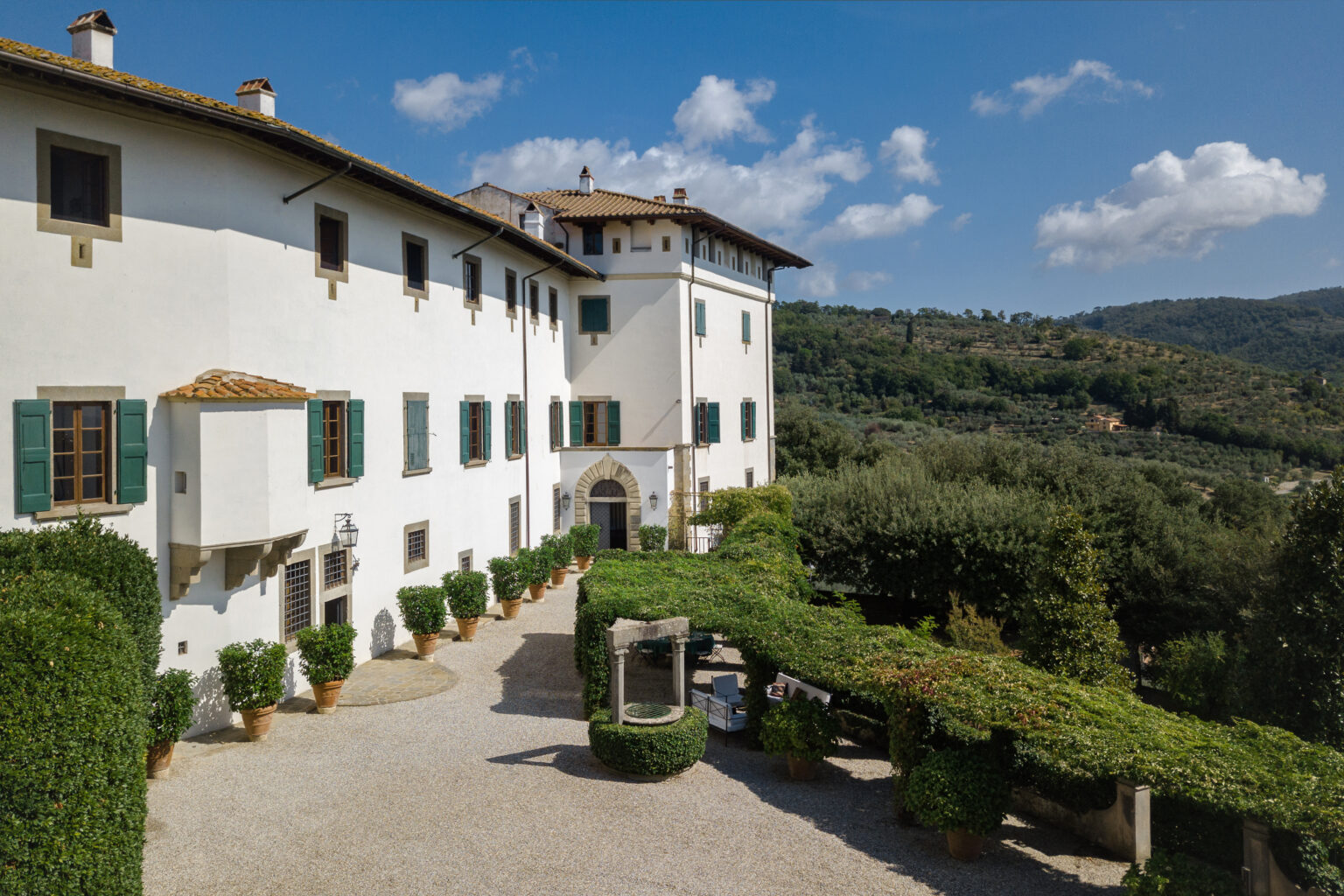 Historic villa surrounded by lush greenery and rolling hills, featuring classic architecture, stone pathways, and vibrant blue skies. Ideal for luxury travel and scenic retreats. | Only on SopranoVillas