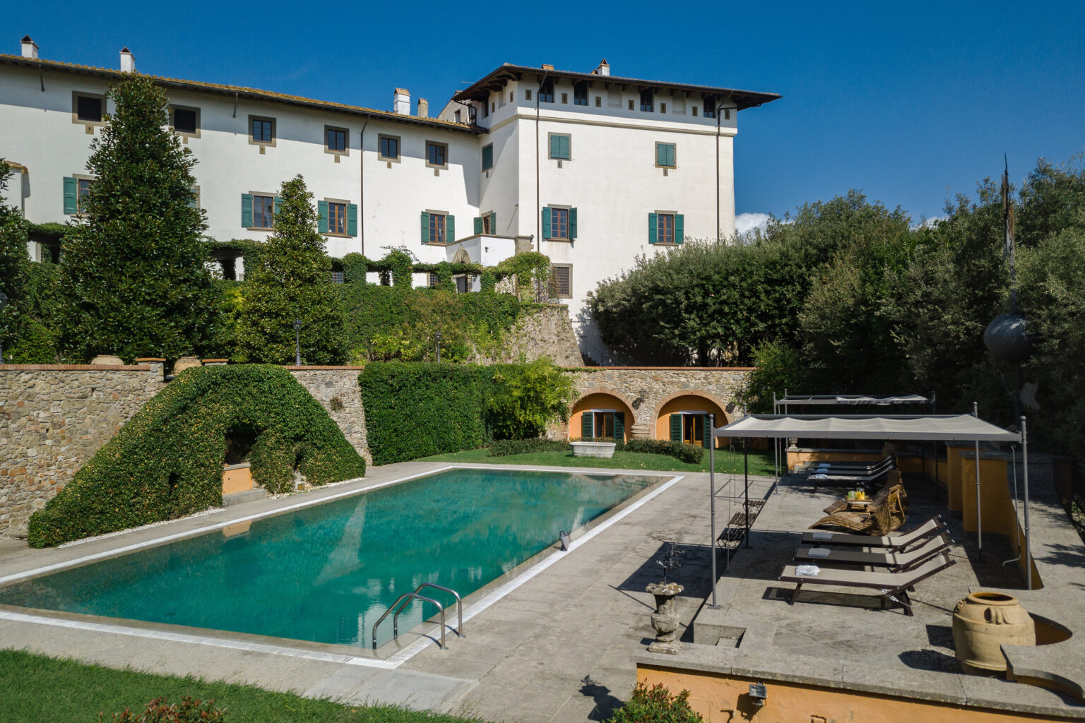 Luxurious villa with a scenic swimming pool surrounded by lush greenery and elegant outdoor seating, set against a clear blue sky. | Only on SopranoVillas