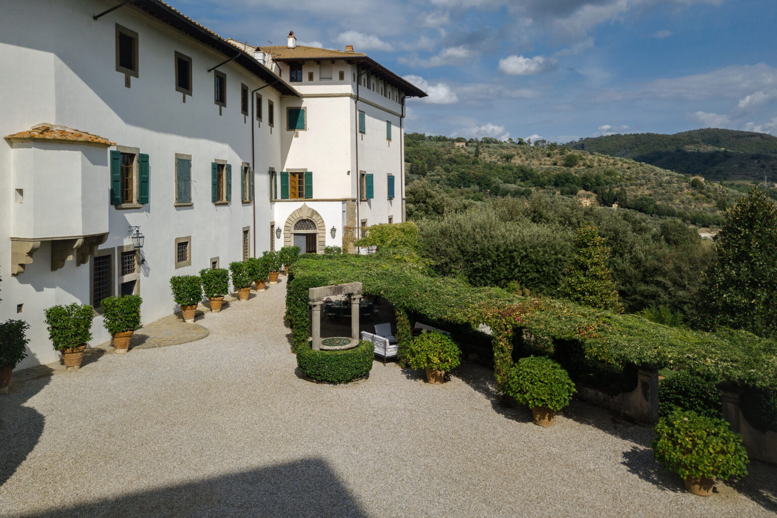 Historic villa with lush gardens and a gravel courtyard, surrounded by rolling hills and olive trees under a clear blue sky. | Only on SopranoVillas