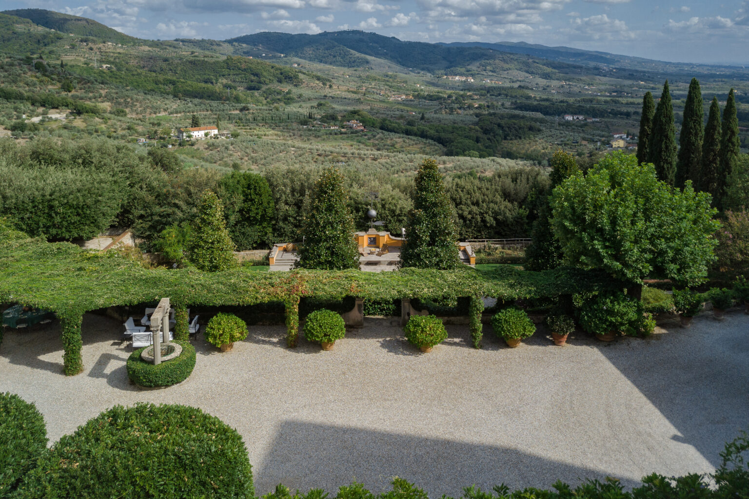 Aerial view of a beautifully landscaped garden featuring manicured hedges, decorative statues, and a gravel pathway, set against a backdrop of rolling hills and olive groves in the countryside. | Only on SopranoVillas