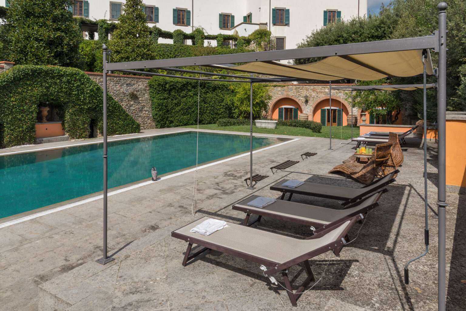 Luxury outdoor pool area featuring sun loungers under a shaded canopy, surrounded by lush greenery and elegant architecture. | Only on SopranoVillas