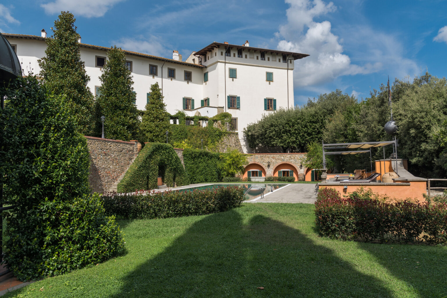 Scenic view of a well-maintained villa surrounded by lush gardens, featuring neatly trimmed hedges and pathways, under a clear blue sky. | Only on SopranoVillas