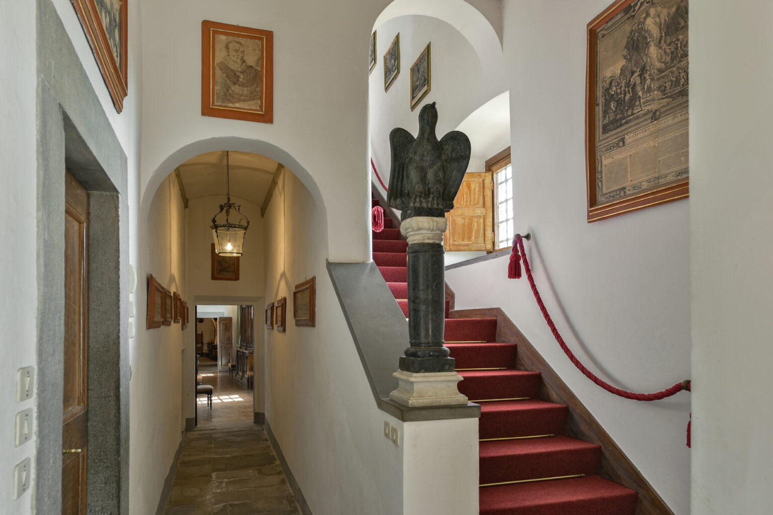 Interior view of a historic building featuring a hallway with arched doorway, red carpeted staircase, and framed artworks on the walls. A decorative sculpture stands at the base of the stairs, enhancing the elegant ambiance. | Only on SopranoVillas