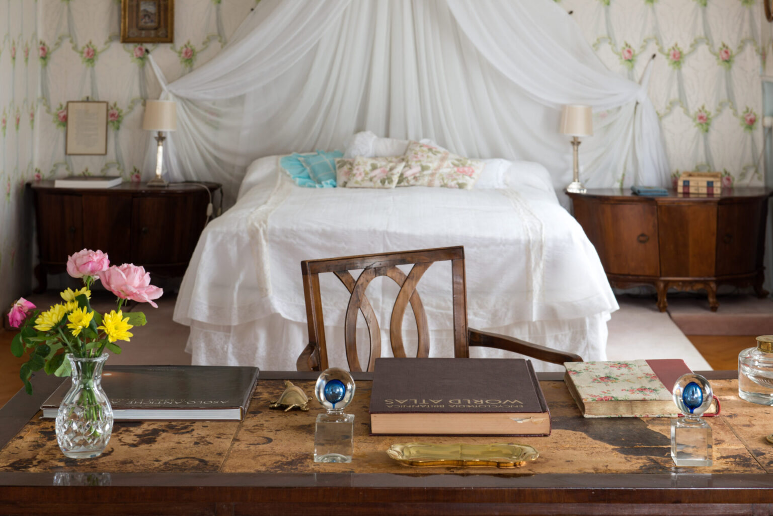 Cozy bedroom interior featuring a wooden desk adorned with books, a crystal vase of flowers, and decorative items. The background showcases a neatly made bed with floral pillows and a soft canopy, complemented by vintage wallpaper and warm lighting. | Only on SopranoVillas