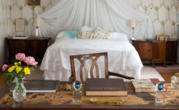 Cozy bedroom interior featuring a wooden desk adorned with books, a crystal vase of flowers, and decorative items. The background showcases a neatly made bed with floral pillows and a soft canopy, complemented by vintage wallpaper and warm lighting.
