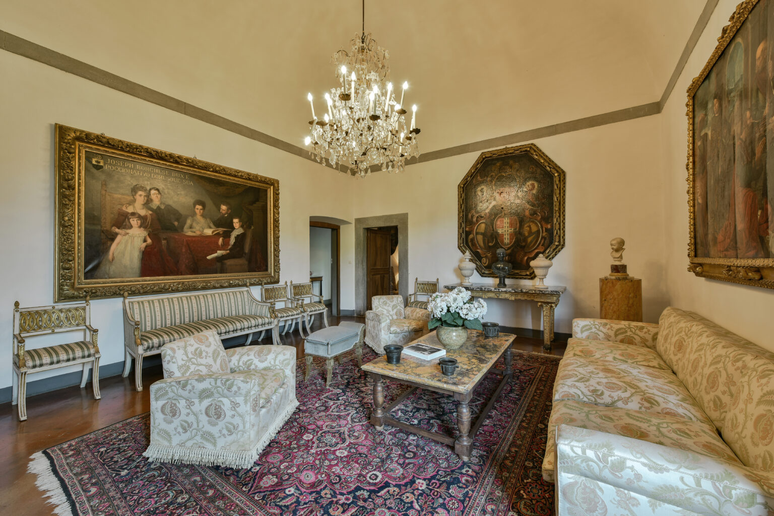 Elegant living room featuring antique furnishings, a crystal chandelier, and classical artwork on the walls. The space includes a patterned area rug, a wooden coffee table adorned with decorative items, and a floral centerpiece, creating a sophisticated atmosphere. | Only on SopranoVillas
