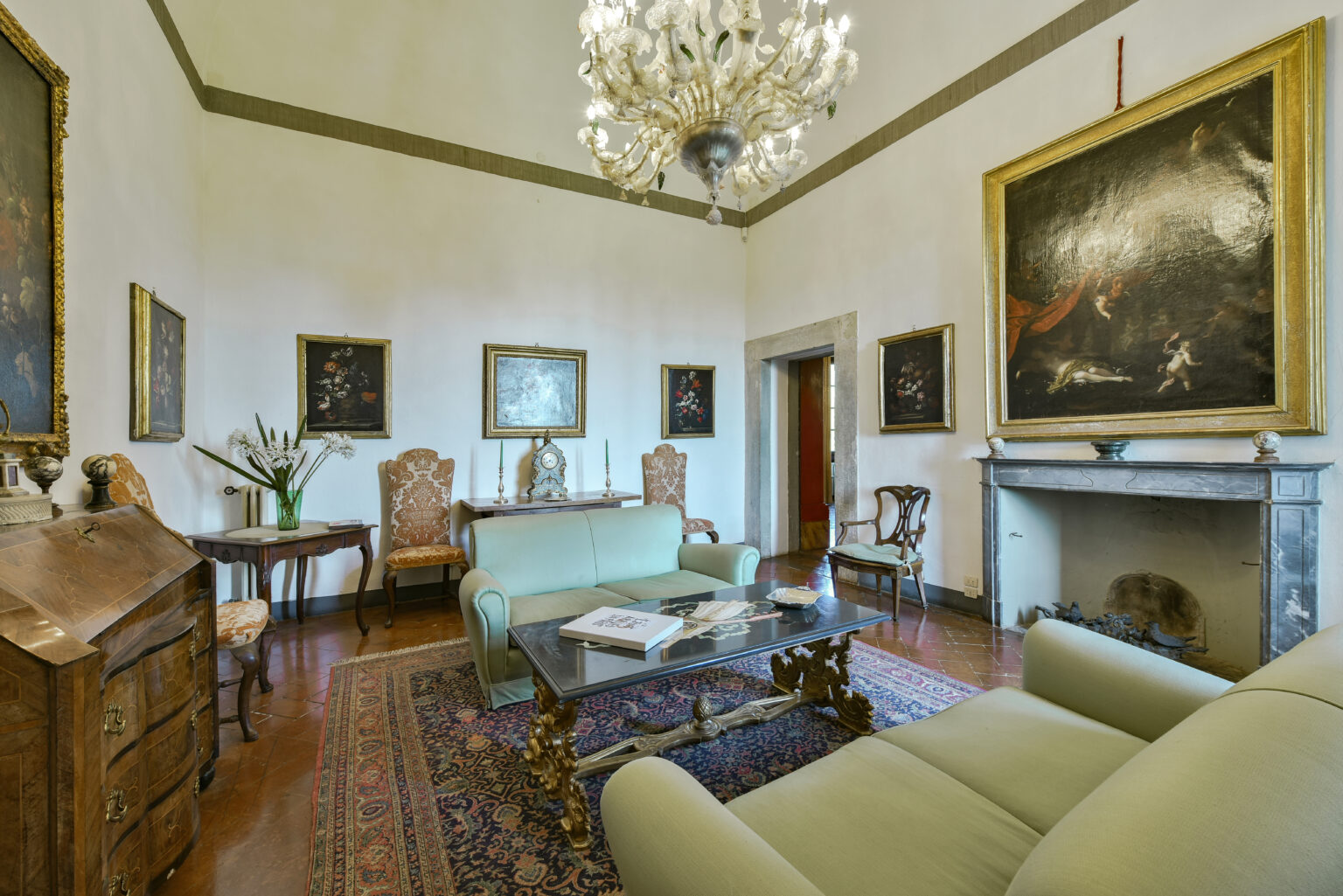 Elegant living room featuring vintage furniture, a large chandelier, and classical artwork. The space includes a green sofa, ornate wooden table, and decorative chairs, all set against a backdrop of neutral walls and a patterned rug. | Only on SopranoVillas