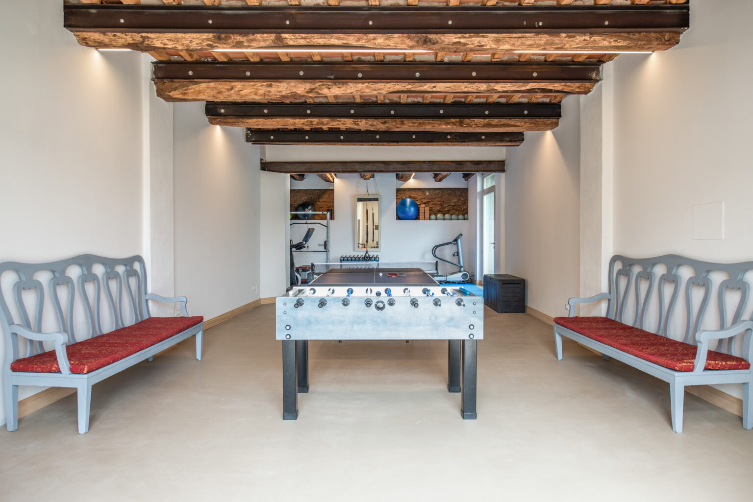Interior view of a recreational room featuring a foosball table, two gray wooden benches with red cushions, and exposed wooden beams. The space is well-lit and includes fitness equipment in the background, creating an inviting atmosphere for relaxation and entertainment. | Only on SopranoVillas