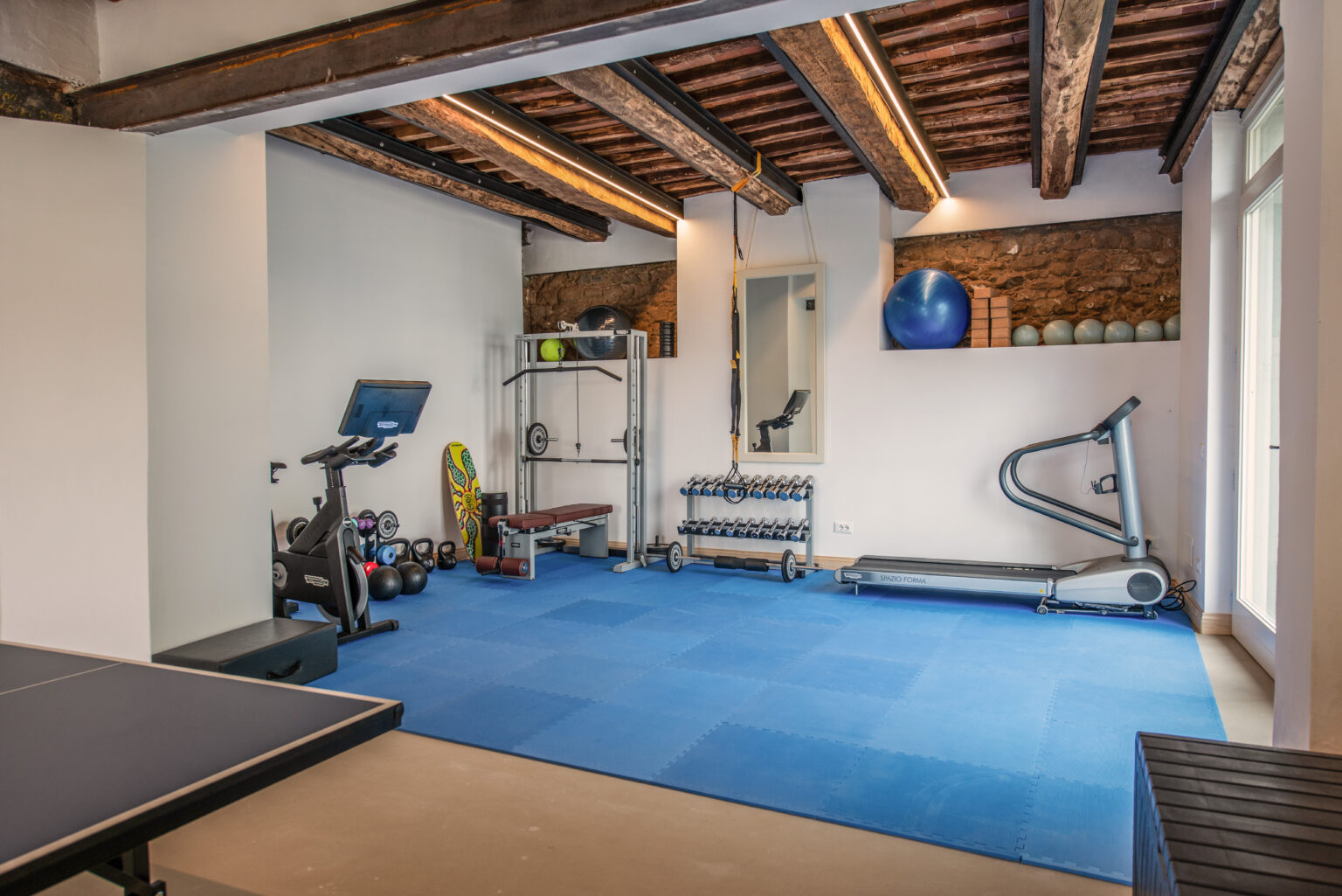 Home gym interior featuring a variety of fitness equipment including a treadmill, weight bench, dumbbells, and a stationary bike, set on a blue rubber floor with wooden beams overhead. | Only on SopranoVillas