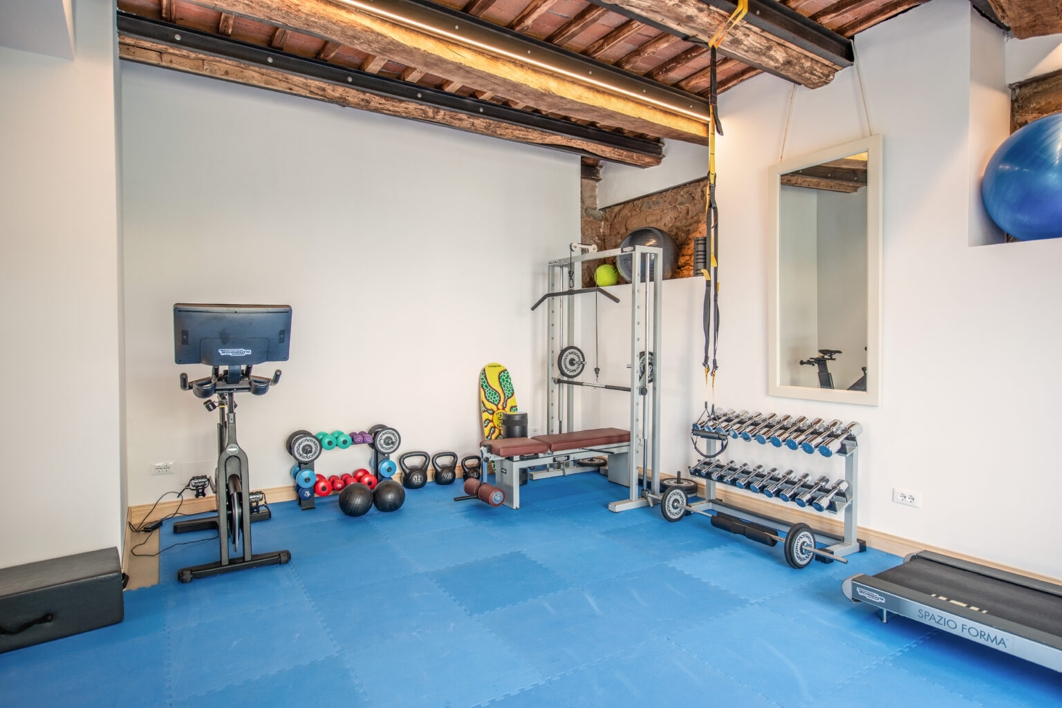 Home gym interior featuring a stationary bike, free weights, kettlebells, a bench press, and exercise equipment on blue rubber flooring, with a large mirror and wooden beams visible in the ceiling. | Only on SopranoVillas