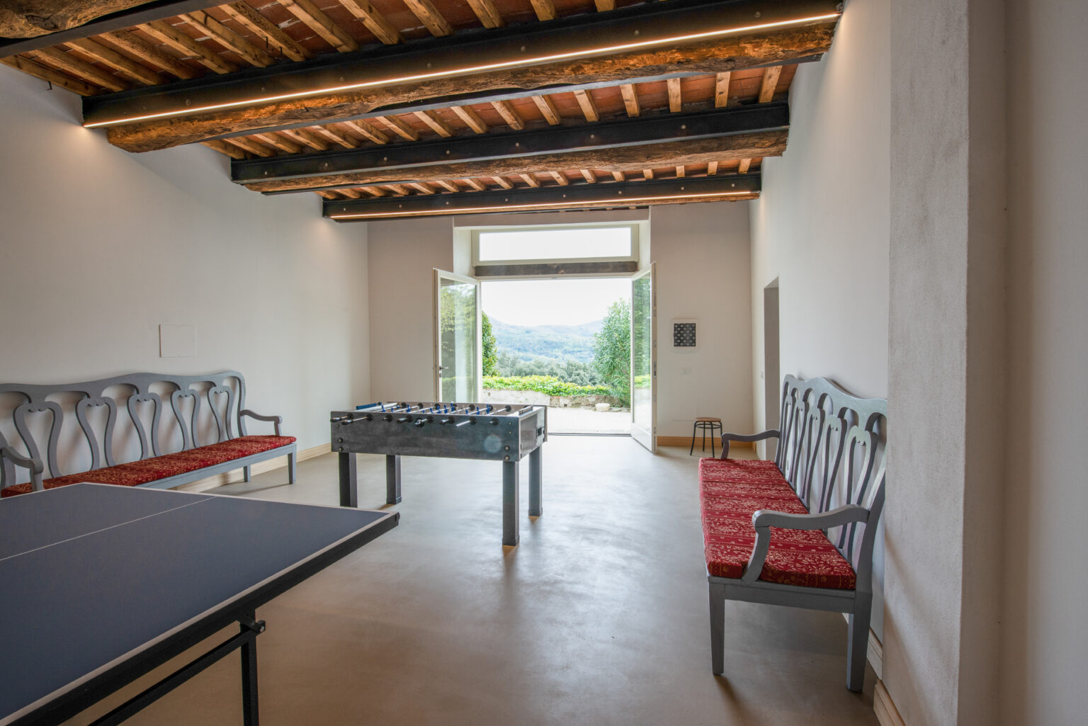Spacious game room featuring a ping pong table and a foosball table, with elegant wooden beams and large open doors offering a scenic view of the outdoors. Stylish gray and red seating enhances the inviting atmosphere. | Only on SopranoVillas