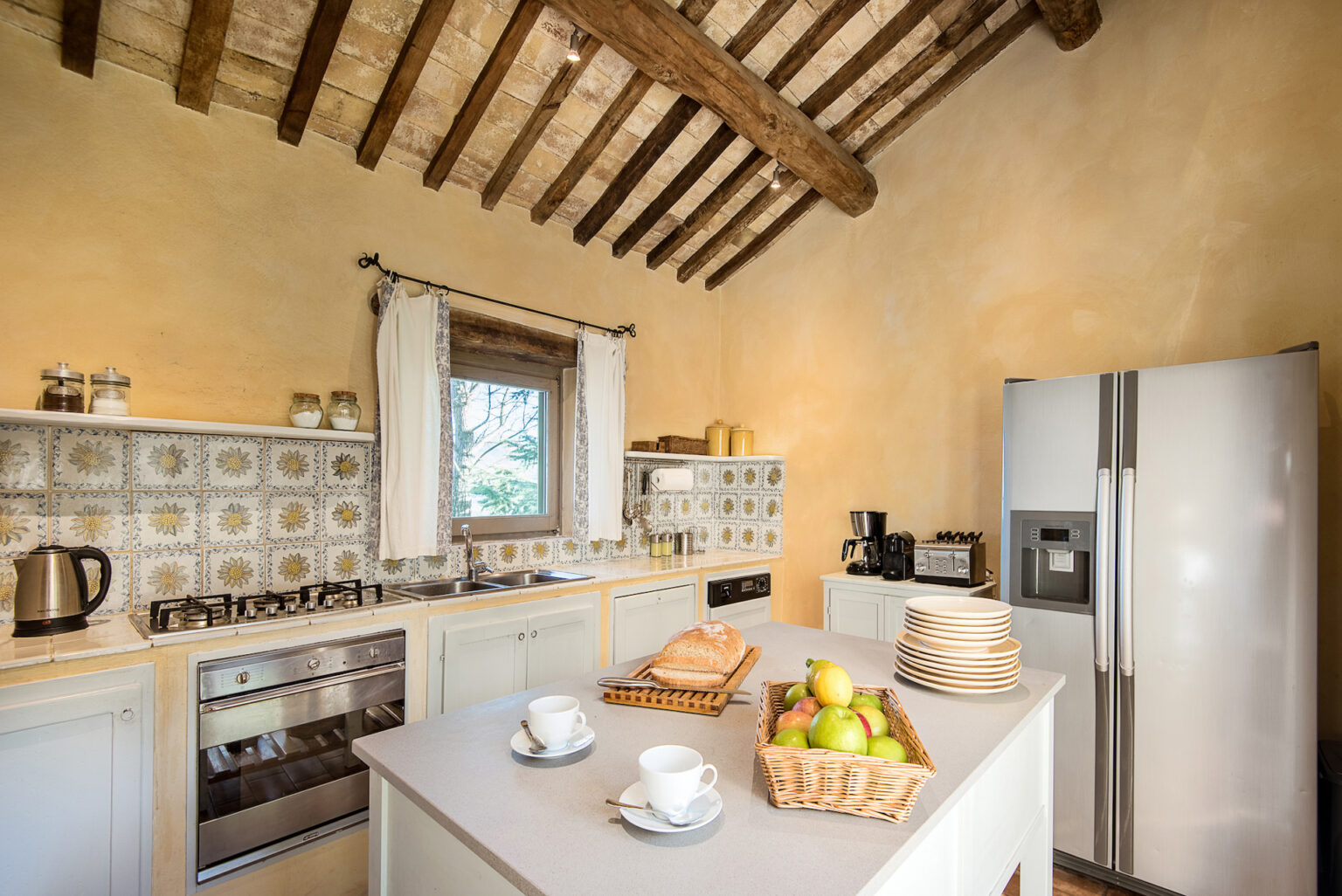 Bright and inviting kitchen featuring rustic wooden beams, tiled backsplash with floral patterns, modern appliances, and a central island with fresh fruit and baked bread. Ideal for culinary enthusiasts and home decor inspiration. | Only on SopranoVillas