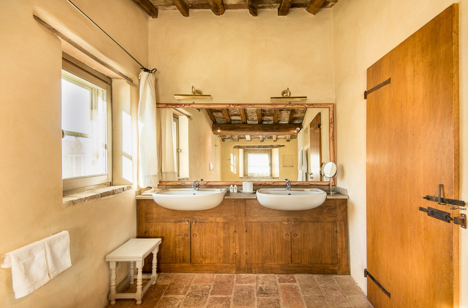 Bright and airy bathroom featuring double sinks, wooden cabinetry, and a large mirror. Natural light streams in through a window, highlighting the rustic decor with exposed wooden beams and terracotta flooring. A small white bench and neatly hung towels add to the inviting atmosphere. | Only on SopranoVillas