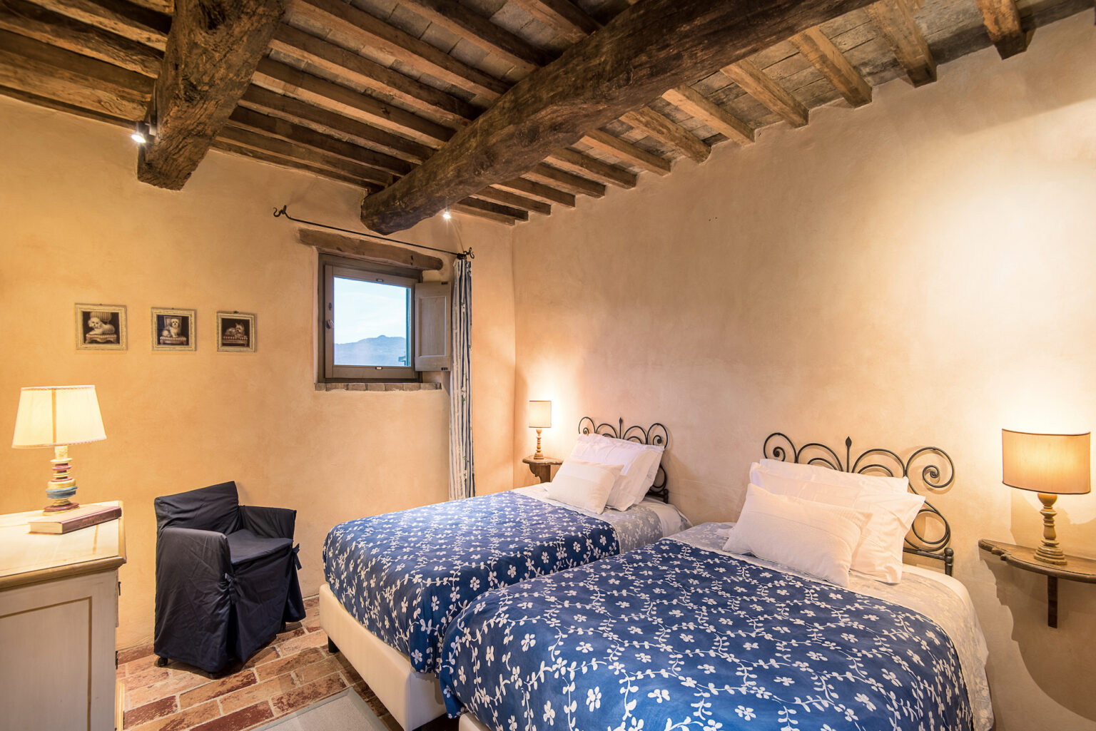 Cozy bedroom featuring two twin beds with blue floral bedspreads, wooden beams on the ceiling, and warm lighting. A window offers a scenic view, while a bedside lamp and decorative chair enhance the inviting atmosphere. Ideal for a relaxing stay. | Only on SopranoVillas