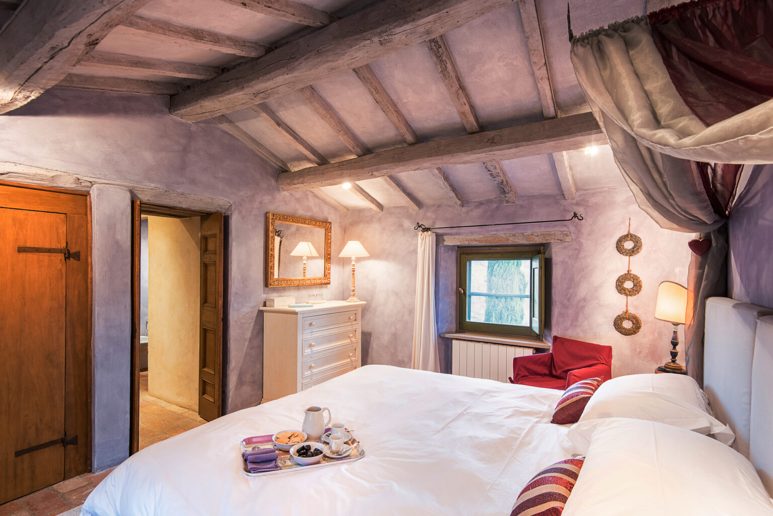 Cozy bedroom with rustic wooden beams, featuring a comfortable bed, a dresser, and a window with natural light. Decorated in soft purple tones, the room includes a red armchair and a small table with refreshments. | Only on SopranoVillas