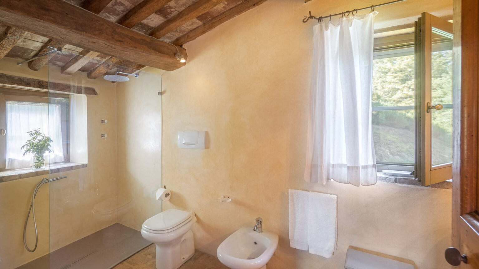 Modern bathroom with rustic charm featuring a bidet, toilet, and large window with natural light. The walls are painted in warm tones, complemented by exposed wooden beams and a sheer white curtain. | Only on SopranoVillas