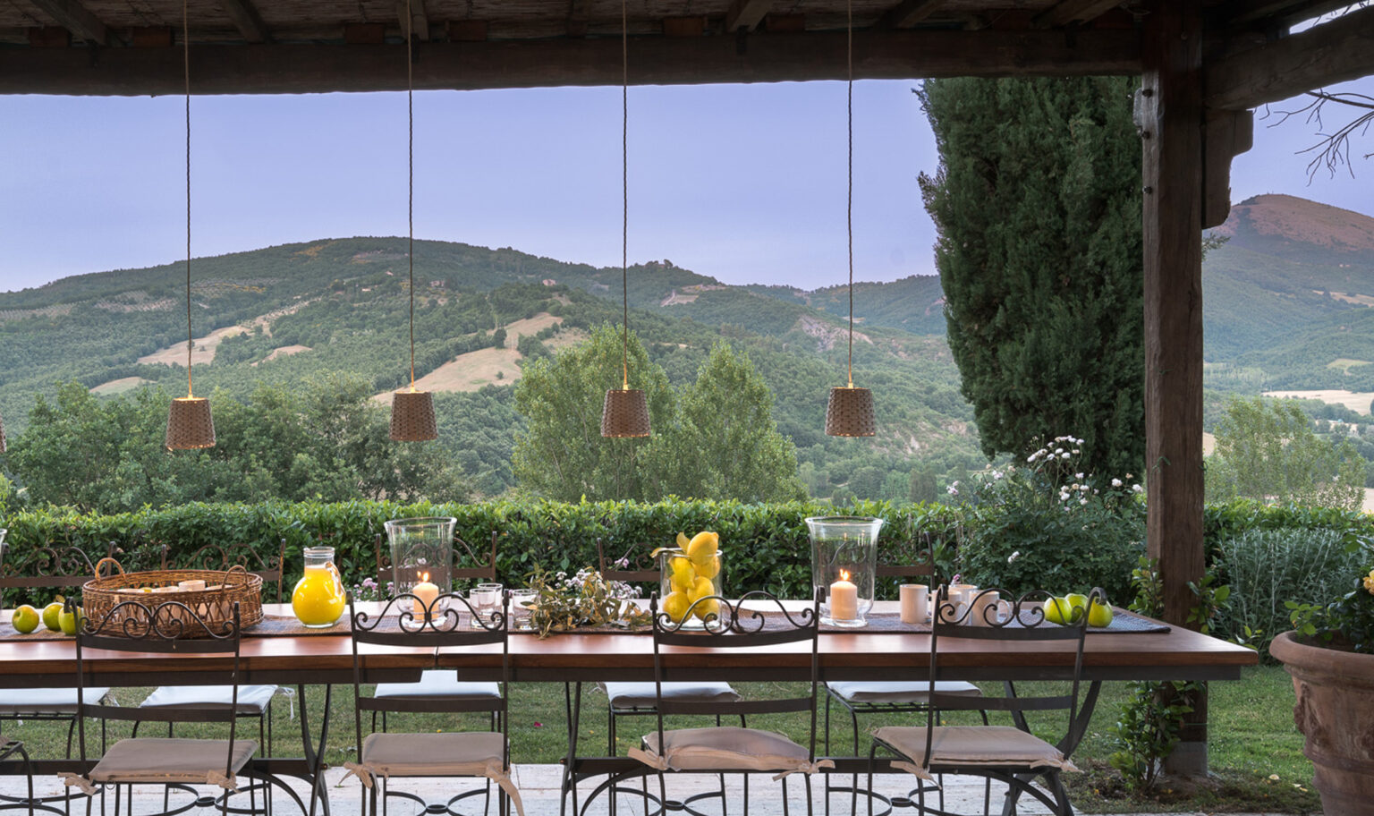 Outdoor dining area with a wooden table set for a meal, surrounded by lush green hills and trees, featuring decorative hanging lanterns and a scenic view of the landscape. | Only on SopranoVillas