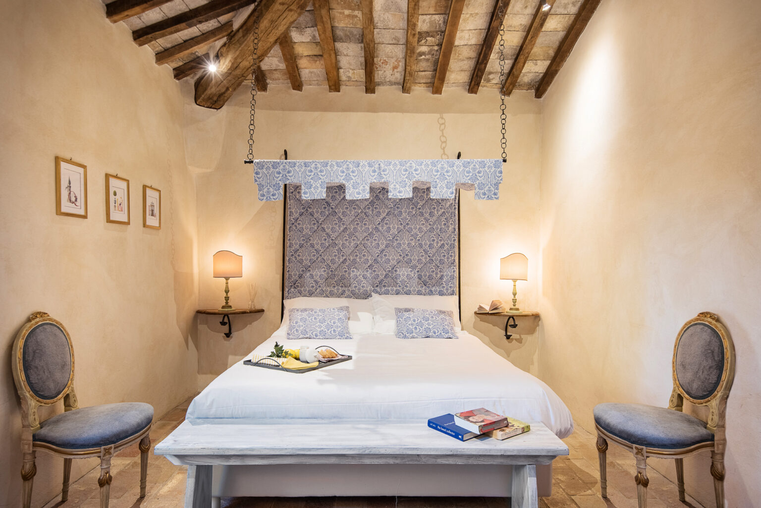 Cozy bedroom featuring a double bed with a decorative headboard, complemented by soft lighting from two bedside lamps. The rustic charm is enhanced by exposed wooden beams and elegant chairs, creating a serene and inviting atmosphere. | Only on SopranoVillas
