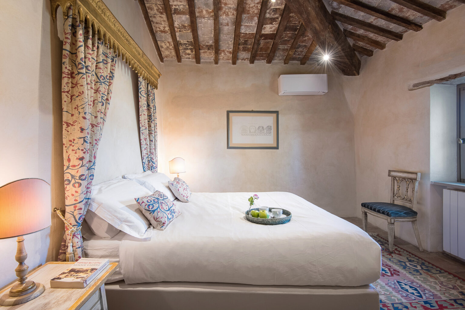 Cozy bedroom featuring a king-size bed with decorative pillows, rustic wooden beams, and elegant curtains. The room includes a bedside lamp, a small tray with greenery, and a vintage chair, creating a warm and inviting atmosphere. | Only on SopranoVillas