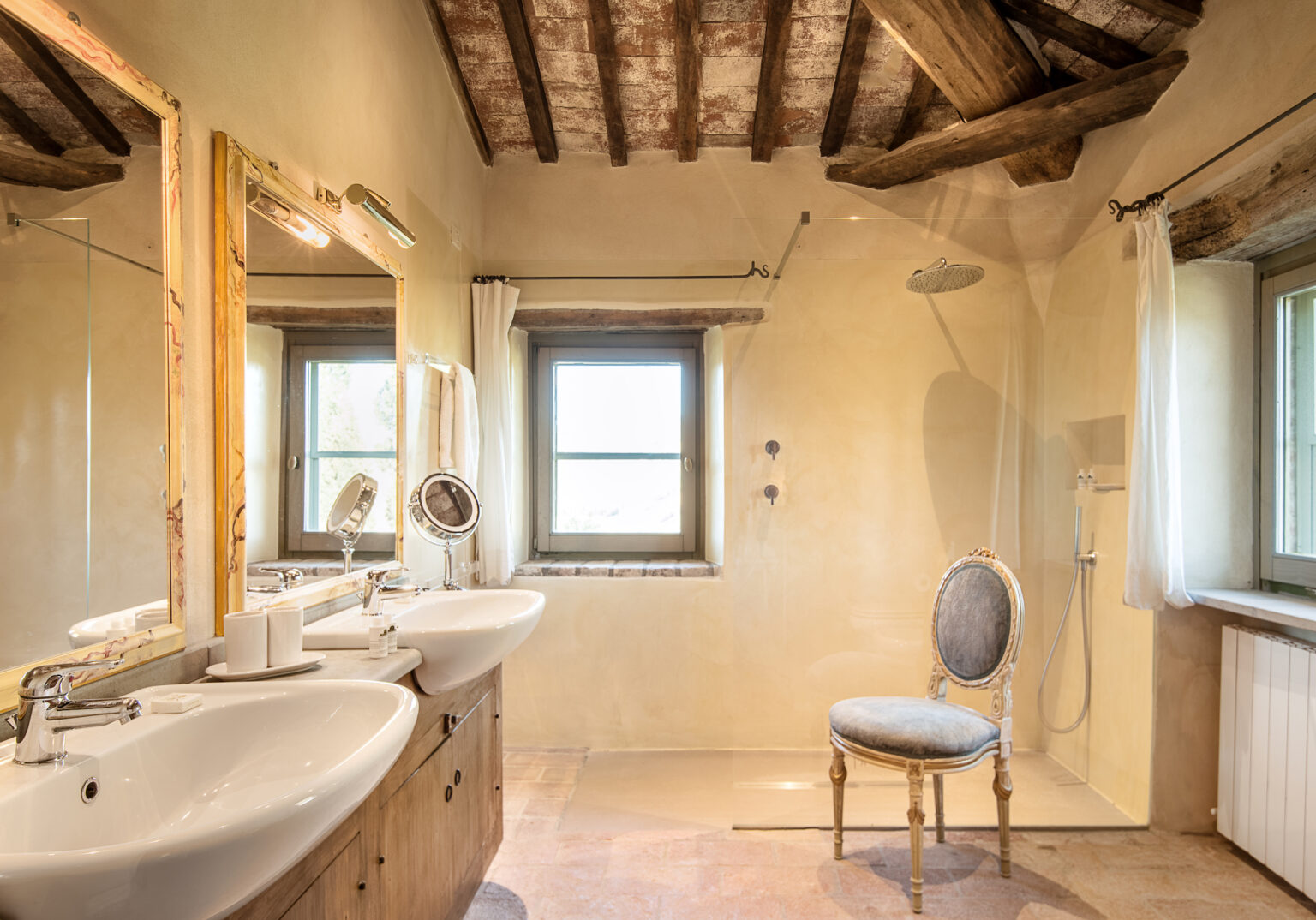 Modern bathroom interior featuring double sinks, a stylish mirror, and a spacious shower area with natural light streaming through a window. The rustic design includes exposed wooden beams and a vintage chair, creating a blend of elegance and comfort. Ideal for home decor inspiration. | Only on SopranoVillas