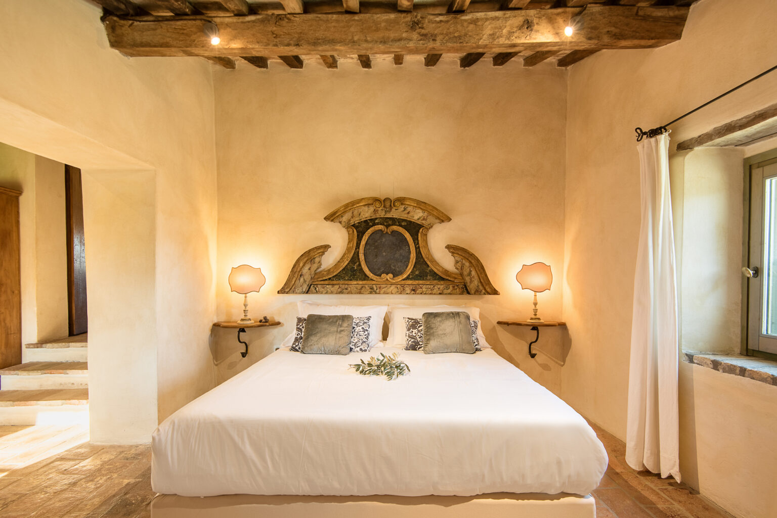 Cozy and elegantly designed bedroom featuring a large bed with decorative headboard, soft lighting from bedside lamps, and rustic wooden beams in the ceiling. The warm color palette and natural decor elements create a serene and inviting atmosphere. | Only on SopranoVillas