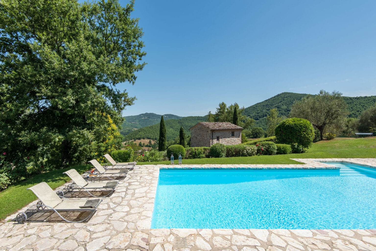 Scenic view of a tranquil poolside area with lounge chairs, surrounded by lush greenery and rolling hills under a clear blue sky. Ideal for relaxation and outdoor leisure. | Only on SopranoVillas