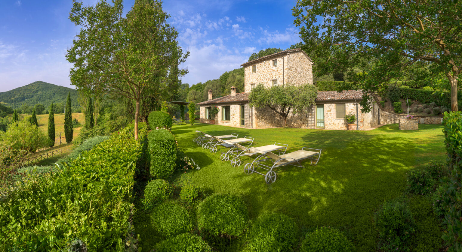 Scenic view of a rustic stone villa surrounded by lush greenery and manicured gardens, featuring sun loungers and cypress trees against a clear blue sky in a serene countryside setting. | Only on SopranoVillas