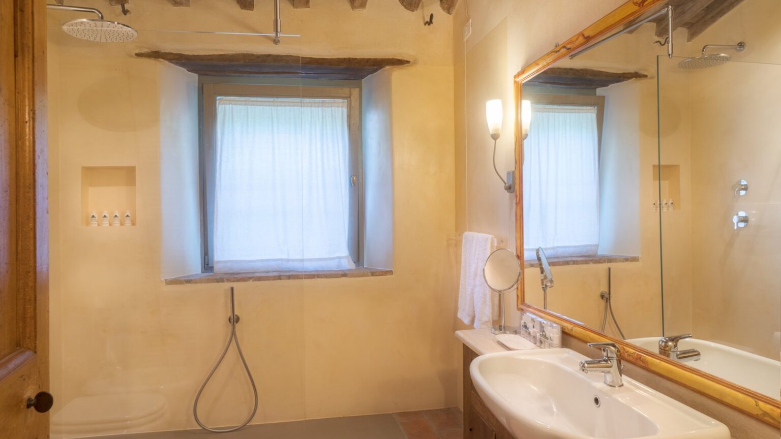 Bright and airy bathroom featuring a large window with sheer curtains, a modern sink with a mirror, and a shower area. The rustic charm is highlighted by wooden beams and warm wall tones, creating a cozy atmosphere. | Only on SopranoVillas