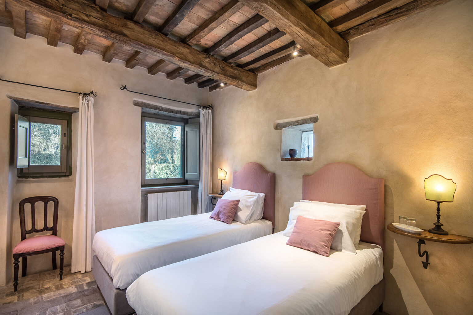 Cozy bedroom featuring two twin beds with soft pink headboards, a wooden ceiling with exposed beams, and natural light streaming through two windows. The room includes a decorative chair and a vintage lamp, creating a warm and inviting atmosphere. Ideal for a relaxing getaway. | Only on SopranoVillas