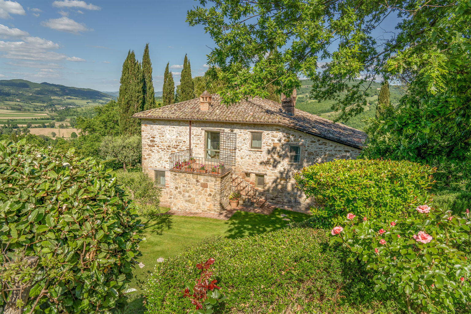 Charming stone cottage surrounded by lush greenery and blooming flowers, set against a picturesque landscape of rolling hills and cypress trees in Tuscany. Ideal for nature lovers and those seeking a tranquil getaway. | Only on SopranoVillas