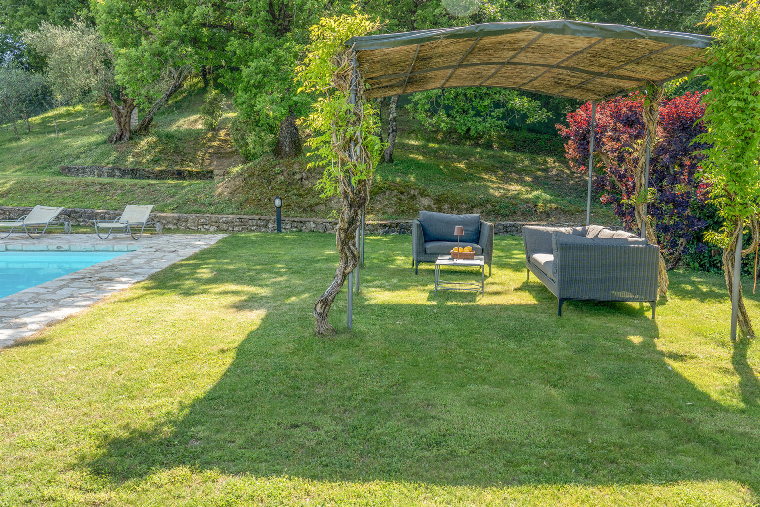 Cozy outdoor seating area under a shaded pergola, surrounded by lush greenery and a swimming pool, perfect for relaxation and leisure in a tranquil garden setting. | Only on SopranoVillas