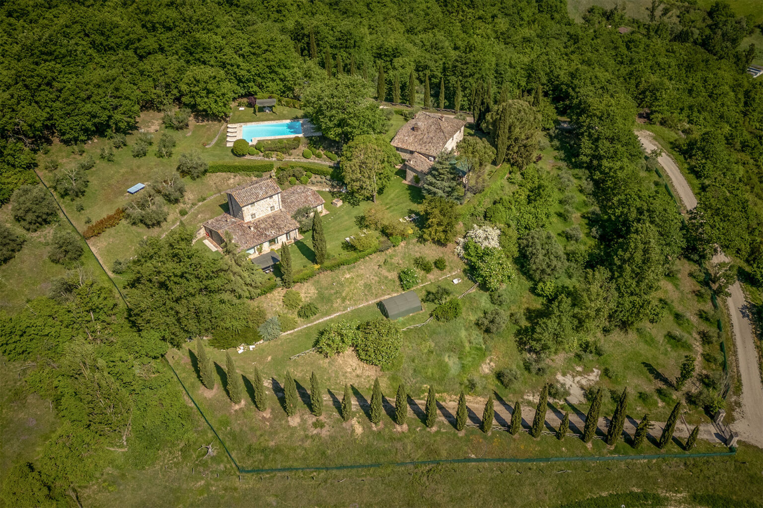 Aerial view of a luxurious villa surrounded by lush greenery, featuring a swimming pool, landscaped gardens, and a row of cypress trees, showcasing a serene countryside setting. | Only on SopranoVillas